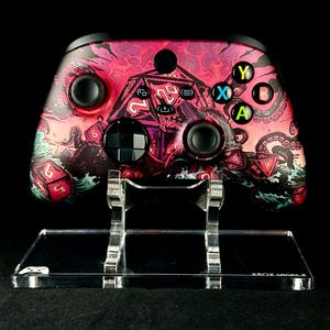 Peut inclure: Une manette Xbox Series X personnalisée avec un motif nautique comprenant des dés et une pieuvre. La manette a des boutons noirs et un support transparent. Le fond est noir.