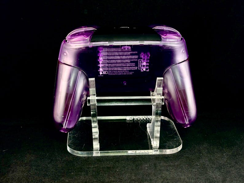 Op de afbeelding: Een transparante paarse gamecontroller op een heldere acryl standaard. De controller heeft een zwarte bovenkant en rust op een heldere, rechthoekige basis. De achtergrond is zwart.