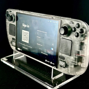 Può includere: Una console di gioco portatile trasparente, che mostra i componenti interni, su un supporto in acrilico trasparente. Lo schermo mostra una pagina di accesso. Il supporto è etichettato "STEAM DECK".