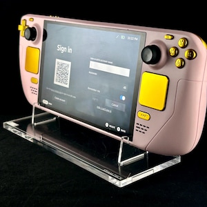 Peut inclure: Console de jeu portable rose et jaune, affichant un écran "Sign in". L'appareil est posé sur un support en acrylique transparent. L'écran montre un code QR et des champs d'informations de compte.
