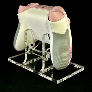 May include: A white and pink gaming controller rests on a clear acrylic stand. The controller has gray grips and pink accents. The stand is rectangular with a clear, reflective surface.