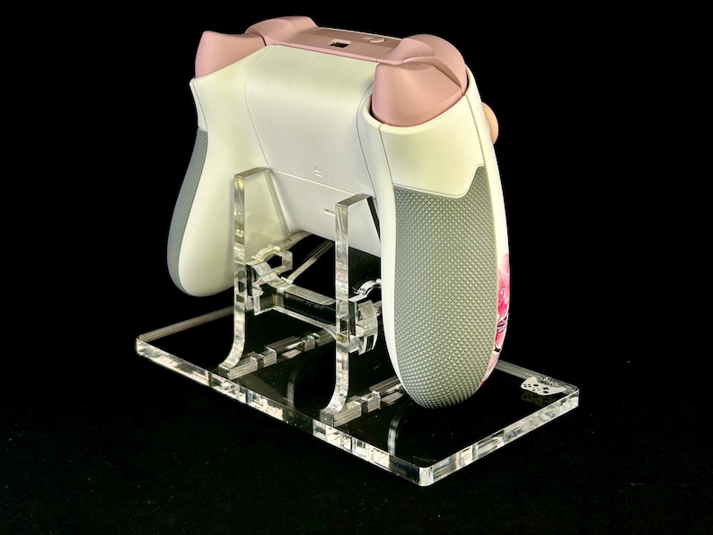 May include: A white and pink gaming controller rests on a clear acrylic stand. The controller has gray grips and pink accents. The stand is rectangular with a clear, reflective surface.