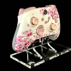 May include: A white and pink Xbox controller featuring a cherry blossom design. The controller has pink buttons and accents, and is displayed on a clear acrylic stand against a black background.