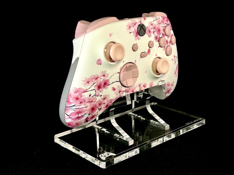 May include: A white and pink Xbox controller featuring a cherry blossom design. The controller has pink buttons and accents, and is displayed on a clear acrylic stand against a black background.