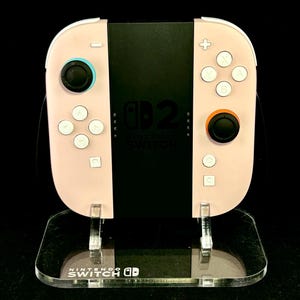 May include: A light pink Nintendo Switch Joy-Con controller with white buttons and black joysticks. The controller is on a clear acrylic stand with the Nintendo Switch logo.