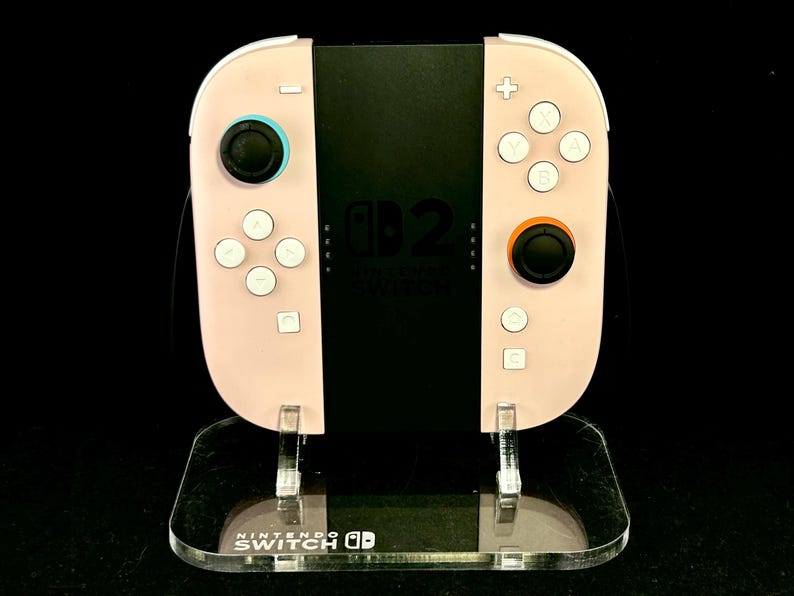 May include: A light pink Nintendo Switch Joy-Con controller with white buttons and black joysticks. The controller is on a clear acrylic stand with the Nintendo Switch logo.