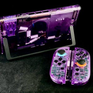 Peut inclure: Une console de jeu violette transparente avec un ensemble de contrôleurs assortis. La console est soutenue par un support noir, révélant ses composants internes. Les contrôleurs sont divisés, avec des boutons colorés.