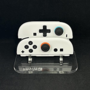 May include: Two white Nintendo Switch Joy-Con controllers stacked on a clear acrylic stand. The controllers have black, orange, and blue buttons. The stand has the words "NINTENDO SWITCH" printed on it.