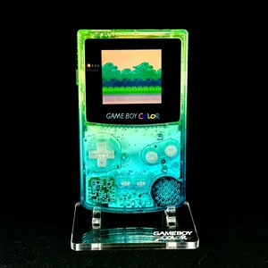 May include: A transparent green and blue Game Boy Color handheld console, displaying a pixelated landscape on its screen. The console is mounted on a clear acrylic stand, with the words "GAME BOY COLOR" visible. The background is black.