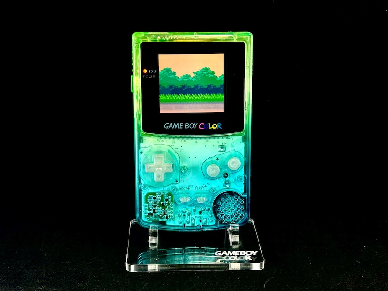 May include: A transparent green and blue Game Boy Color handheld console, displaying a pixelated landscape on its screen. The console is mounted on a clear acrylic stand, with the words "GAME BOY COLOR" visible. The background is black.