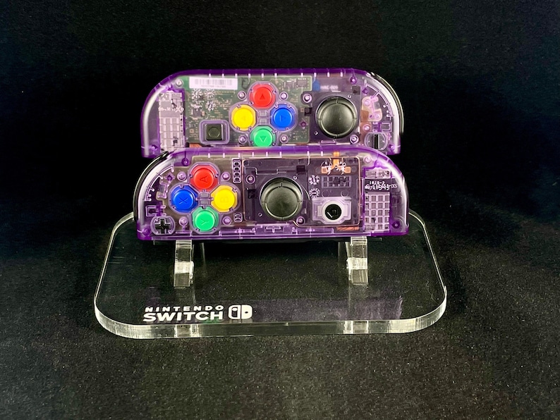 Pu&ograve; includere: Due controller Nintendo Switch Joy-Con trasparenti viola con pulsanti colorati sono esposti su un supporto in acrilico trasparente con la scritta "NINTENDO SWITCH" stampata sulla base.