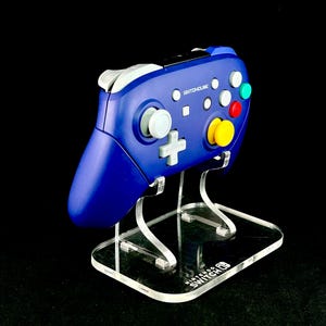 K&ouml;nnte beinhalten: Ein blauer SwitchHouse-Controller mit wei&szlig;en, roten, gelben und gr&uuml;nen Kn&ouml;pfen, pr&auml;sentiert auf einem transparenten Acrylst&auml;nder. Der St&auml;nder hat ein Nintendo Switch-Logo auf der Basis. Der Controller hat ein klassisches Design.