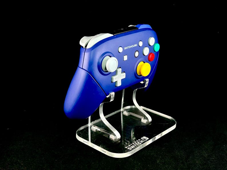 K&ouml;nnte beinhalten: Ein blauer SwitchHouse-Controller mit wei&szlig;en, roten, gelben und gr&uuml;nen Kn&ouml;pfen, pr&auml;sentiert auf einem transparenten Acrylst&auml;nder. Der St&auml;nder hat ein Nintendo Switch-Logo auf der Basis. Der Controller hat ein klassisches Design.