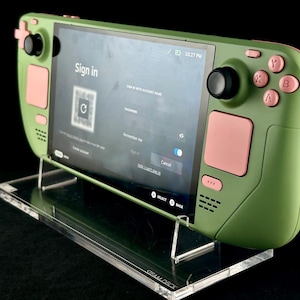 Könnte beinhalten: Eine grüne Handheld-Spielekonsole mit rosa Akzenten, die einen Anmeldebildschirm anzeigt. Die Konsole steht auf einem transparenten Acrylständer. Der Bildschirm zeigt einen QR-Code und Anmeldeoptionen. Die Konsole hat Tasten und Joysticks.