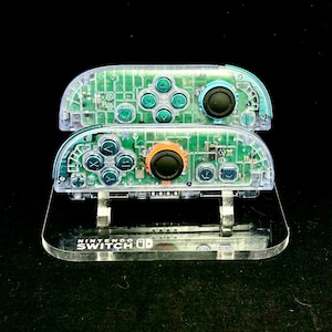 May include: Two clear Nintendo Switch Joy-Con controllers with visible internal circuitry. The controllers have teal buttons and black and orange joysticks. They rest on a clear acrylic stand with the Nintendo Switch logo.