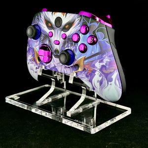 May include: A custom video game controller with a wolf design in purple, white, and blue, resting on a clear acrylic stand. The controller features purple buttons and blue thumbsticks. The stand is clear and rectangular.