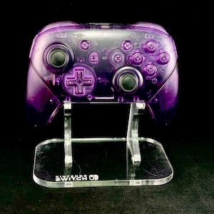 Op de afbeelding: Een transparante paarse Nintendo Switch Pro Controller op een heldere acryl standaard. De controller heeft zwarte knoppen en een D-pad. De standaard heeft de tekst "Nintendo Switch" op de basis.