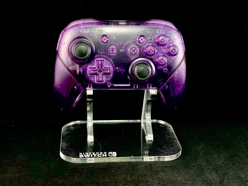 Op de afbeelding: Een transparante paarse Nintendo Switch Pro Controller op een heldere acryl standaard. De controller heeft zwarte knoppen en een D-pad. De standaard heeft de tekst "Nintendo Switch" op de basis.