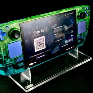 May include: A handheld gaming console with a translucent neon green shell and blue buttons, joysticks, and trackpads. The screen displays a sign-in interface with a QR code, the text Sign in, and various login fields. It rests on a clear acrylic stand.