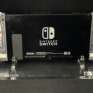 May include: A black Nintendo Switch console with a clear plastic stand. The console has a white Nintendo Switch logo and the words "Nintendo Switch" printed on the back.