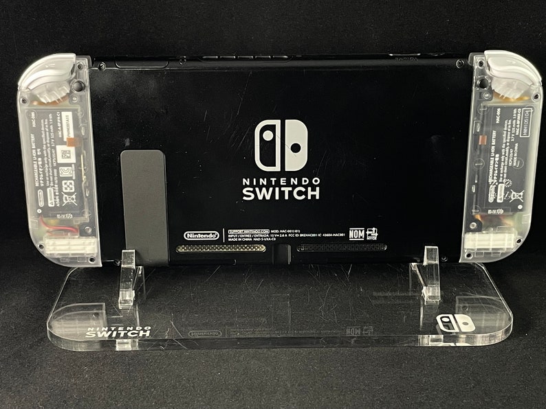 May include: A black Nintendo Switch console with a clear plastic stand. The console has a white Nintendo Switch logo and the words "Nintendo Switch" printed on the back.