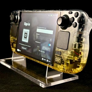 May include: A transparent handheld gaming device with a black screen displaying a sign-in page. The device has a clear yellow casing and black buttons. It is sitting on a clear acrylic stand.