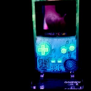 May include: A glowing, transparent Game Boy Color handheld console. The device has a neon green and blue color scheme, with the screen displaying a pink and purple image. The text "GAME BOY COLOR" is visible on the device.