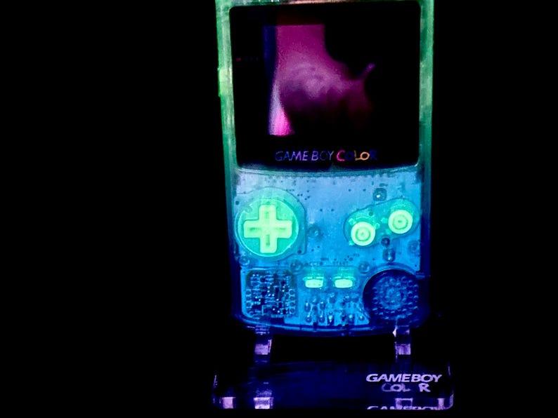 May include: A glowing, transparent Game Boy Color handheld console. The device has a neon green and blue color scheme, with the screen displaying a pink and purple image. The text "GAME BOY COLOR" is visible on the device.