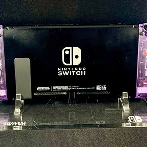 Pu&ograve; includere: Una console Nintendo Switch nera con controller Joy-Con viola su un supporto in acrilico trasparente. La console ha il logo Nintendo Switch e le parole "Nintendo Switch" stampate sul retro.