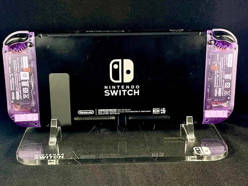 Pu&ograve; includere: Una console Nintendo Switch nera con controller Joy-Con viola su un supporto in acrilico trasparente. La console ha il logo Nintendo Switch e le parole "Nintendo Switch" stampate sul retro.