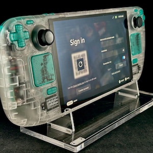 Könnte beinhalten: Ein transparentes Handheld-Gaming-Gerät mit türkisfarbenen Akzenten, das einen Anmeldebildschirm anzeigt. Das Gerät ist auf einem transparenten Acryl-Ständer montiert. Der Bildschirm zeigt einen QR-Code und fordert Kontodaten an.