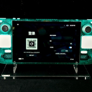 May include: A transparent teal handheld gaming console on a clear stand, displaying a login screen with Chinese characters and a QR code. The console has buttons, joysticks, and a screen. The time is 10:20 PM.