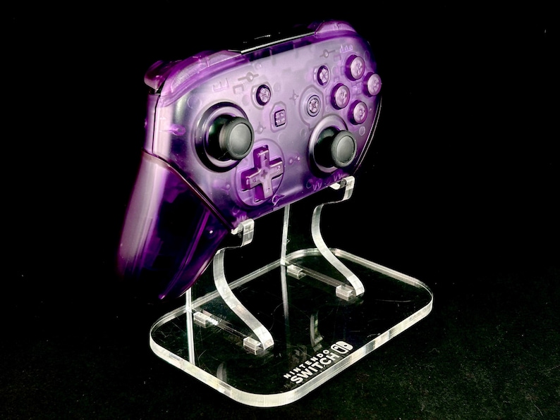 Op de afbeelding: Een transparante paarse Nintendo Switch Pro Controller staat op een heldere acryl standaard. De controller heeft zwarte joysticks en knoppen. De standaard heeft het Nintendo Switch logo op de basis, tegen een zwarte achtergrond.