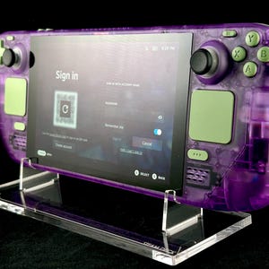 Pode incluir: Um dispositivo de jogos portátil roxo transparente com detalhes verdes, exibindo uma tela de login. O dispositivo é montado em um suporte de acrílico transparente. A tela mostra o texto "Sign in".