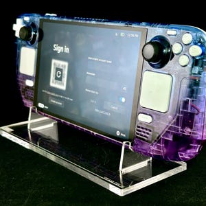 Puede incluir: Una consola de juegos portátil Steam Deck transparente púrpura y azul sobre un soporte acrílico transparente. La pantalla muestra una página de inicio de sesión. La consola tiene joysticks y botones negros. El soporte tiene el texto "STEAM DECK".