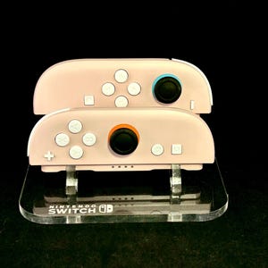 Könnte beinhalten: Zwei rosa Nintendo Switch Joy-Con-Controller sind auf einem transparenten Acrylständer gestapelt. Die Controller haben weiße Knöpfe und schwarze Joysticks mit blauen und orangefarbenen Akzenten. Der Ständer hat das Nintendo Switch-Logo.