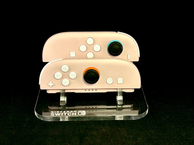 May include: Two pink Nintendo Switch Joy-Con controllers are stacked on a clear acrylic stand. The controllers have white buttons and black joysticks with blue and orange accents. The stand has the Nintendo Switch logo.