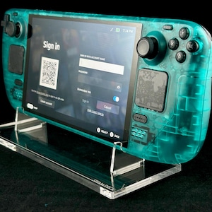 Könnte beinhalten: Eine türkisfarbene Handheld-Spielkonsole mit einem durchsichtigen Kunststoffgehäuse. Die Konsole steht auf einem durchsichtigen Acrylständer. Der Bildschirm zeigt eine Anmeldeseite mit einem QR-Code und einem Textfeld zur Eingabe eines Kontonamens.