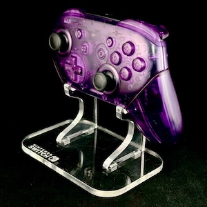 May include: A transparent purple Nintendo Switch Pro Controller on a clear acrylic stand. The controller features black buttons and a directional pad. The stand has the Nintendo Switch logo.