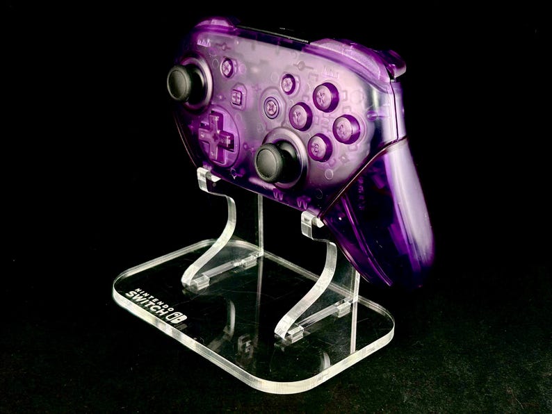 Op de afbeelding: Een transparante paarse Nintendo Switch Pro Controller op een heldere acryl standaard. De controller heeft zwarte knoppen en een D-pad. De standaard heeft het Nintendo Switch logo.