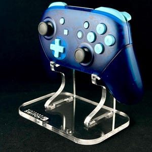 May include: A blue and light blue Nintendo Switch controller on a clear acrylic stand with the text "Nintendo Switch" on the base.