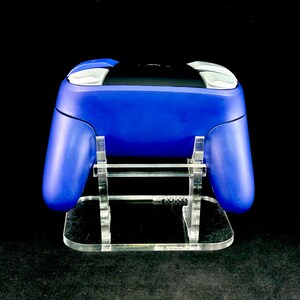 K&ouml;nnte beinhalten: Ein blauer Videospiel-Controller mit wei&szlig;en und silbernen Akzenten, der auf einem transparenten Acryl-St&auml;nder ruht. Der St&auml;nder hat eine rechteckige Basis und tr&auml;gt den Controller, ein modernes Gaming-Zubeh&ouml;r.