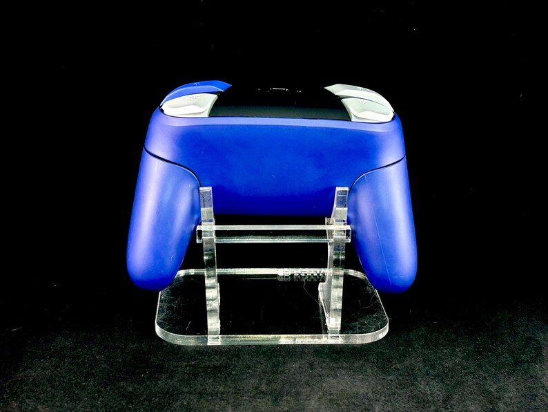 K&ouml;nnte beinhalten: Ein blauer Videospiel-Controller mit wei&szlig;en und silbernen Akzenten, der auf einem transparenten Acryl-St&auml;nder ruht. Der St&auml;nder hat eine rechteckige Basis und tr&auml;gt den Controller, ein modernes Gaming-Zubeh&ouml;r.