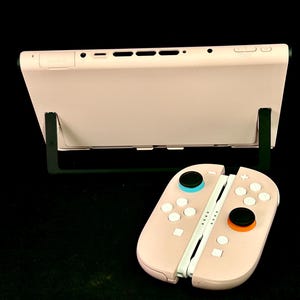 May include: A light pink handheld gaming console with detached controllers. The console is propped up on a black stand. The controllers are light pink with black, blue, and orange buttons. The background is black.