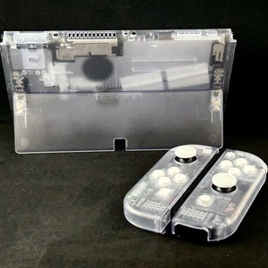Puede incluir: Una carcasa transparente de plástico para una consola Nintendo Switch con dos mandos Joy-Con transparentes de plástico. Los mandos tienen botones blancos.