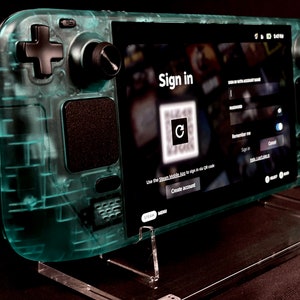 May include: A teal transparent handheld gaming console with black controls and a screen showing a sign-in interface. The screen displays a QR code and prompts for account details. The device is on a clear stand.
