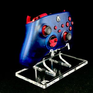 Könnte beinhalten: Ein blau-roter Xbox-Controller ruht auf einem transparenten Acrylständer. Der Controller hat rote Knöpfe und Akzente mit einem blauen Gehäuse. Der Ständer ist rechteckig mit einer transparenten Basis und Halterungen.