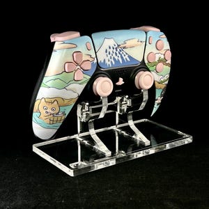 Peut inclure: Une manette de jeu personnalisée avec un design japonais, représentant le Mont Fuji, des fleurs de cerisier et un chat. La manette a des boutons roses et est présentée sur un support en acrylique transparent.