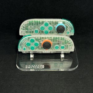 Pode incluir: Dois controles Joy-Con transparentes do Nintendo Switch com circuitos internos visíveis, empilhados em um suporte de acrílico transparente. Os controles têm botões verdes, azuis e laranja. O suporte é marcado com o logotipo do Nintendo Switch.