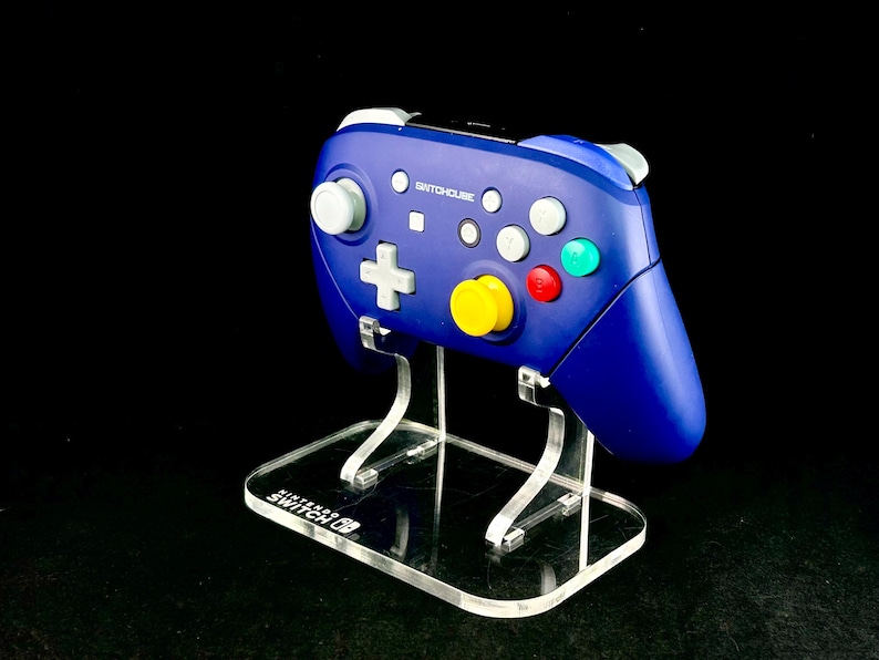 K&ouml;nnte beinhalten: Ein blauer Switch-Controller mit wei&szlig;en, roten, gelben und gr&uuml;nen Kn&ouml;pfen wird auf einem transparenten Acrylst&auml;nder pr&auml;sentiert. Der Controller tr&auml;gt den Text "SWITCHHOLIC". Der St&auml;nder tr&auml;gt den Text "NINTENDO SWITCH".